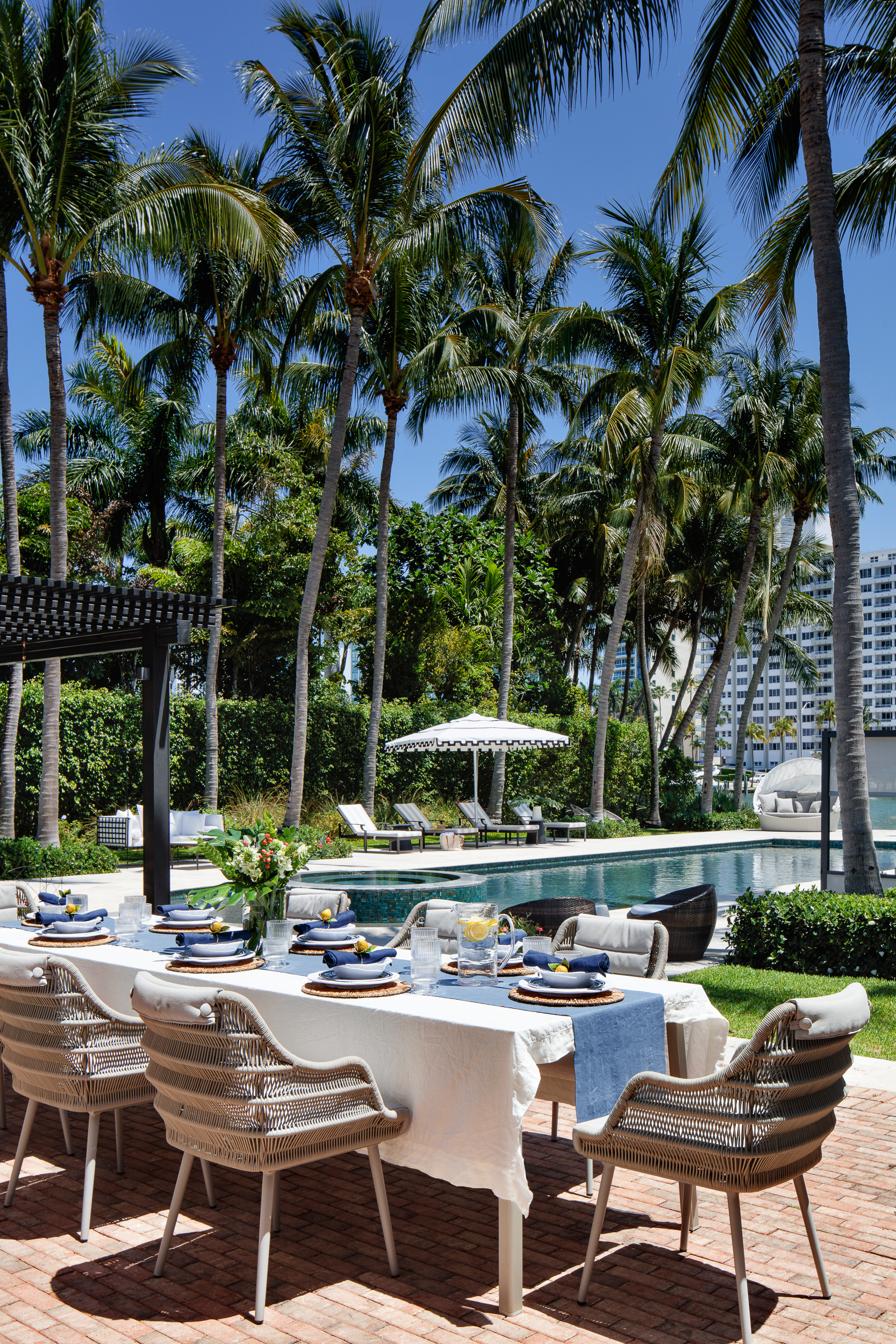 Poolside dining table with striped linens and woven chairs beneath palms.