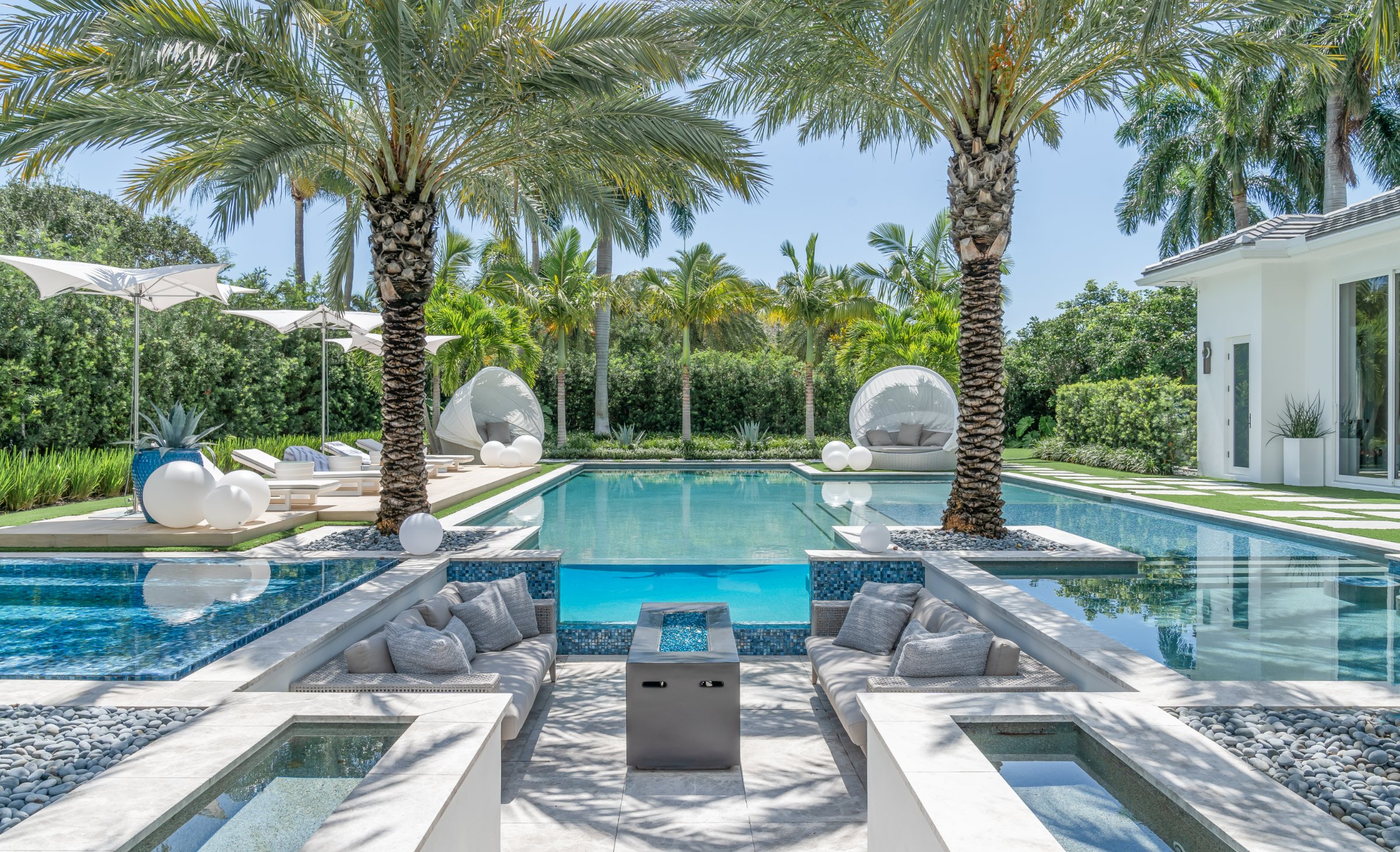 Pool deck in light-colored natural stone flanked by palms and daybeds.